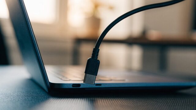 Close-up of a USB-C charger cable plugged into the side port of a dark blue laptop on a dark desk mat