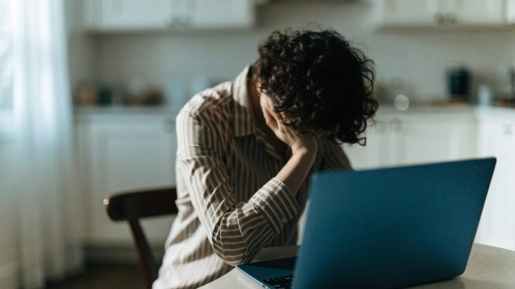 A person resting their forehead on one hand in front of a dark blue laptop on a kitchen table