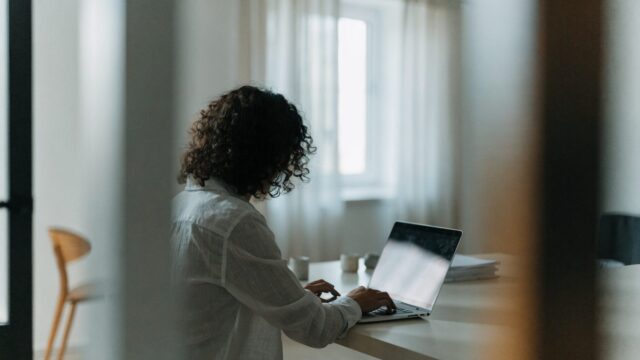 A person seen from behind working on a silver laptop in a bright home office