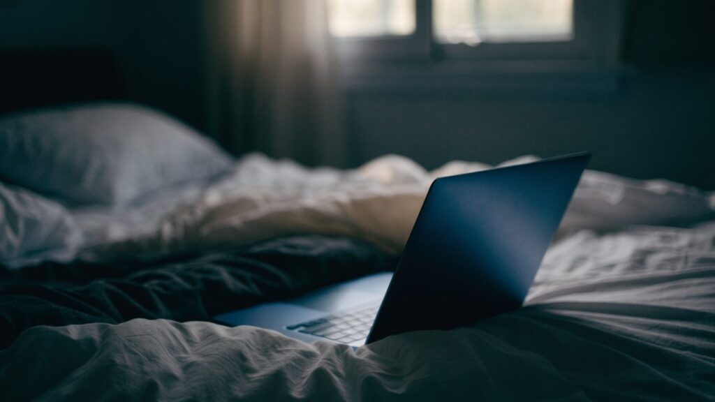 A dark blue laptop glowing softly on a bed in a dark room