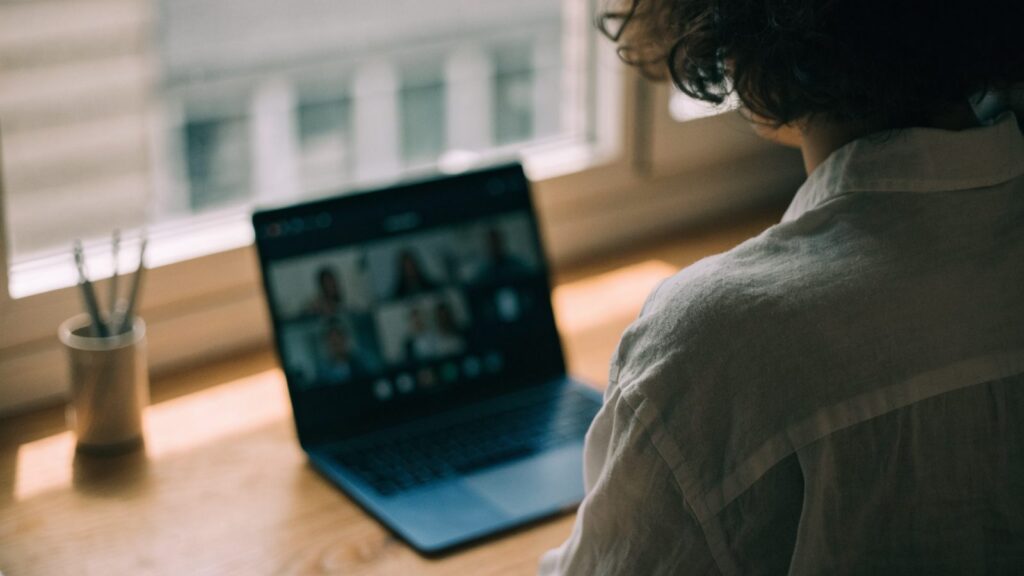 A person seen from behind on a video call on a dark blue laptop on a wooden desk