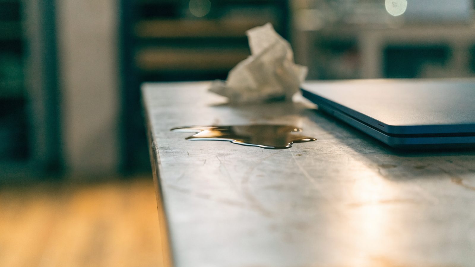 A small puddle of spilled coffee next to a dark blue laptop on a workbench