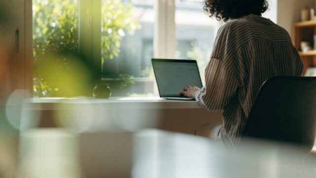 A person seen from behind working on a matte grey laptop in a bright home office