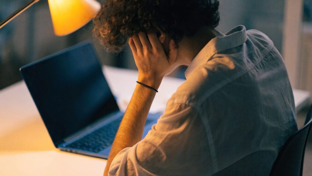 A person resting their forehead on one hand in front of a dark blue laptop on a white desk