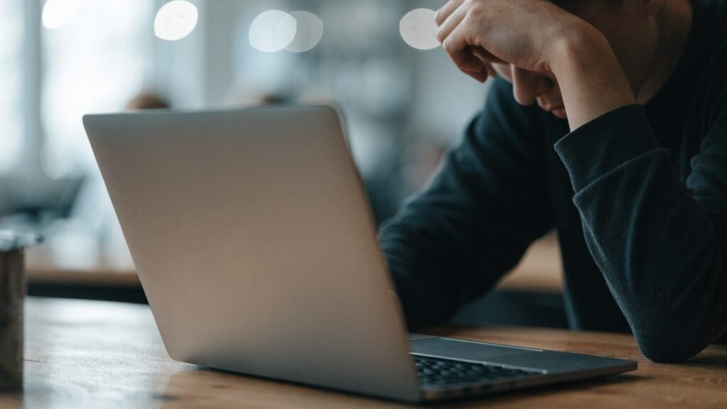 A person resting their forehead on one hand in front of a matte grey laptop on a wooden desk