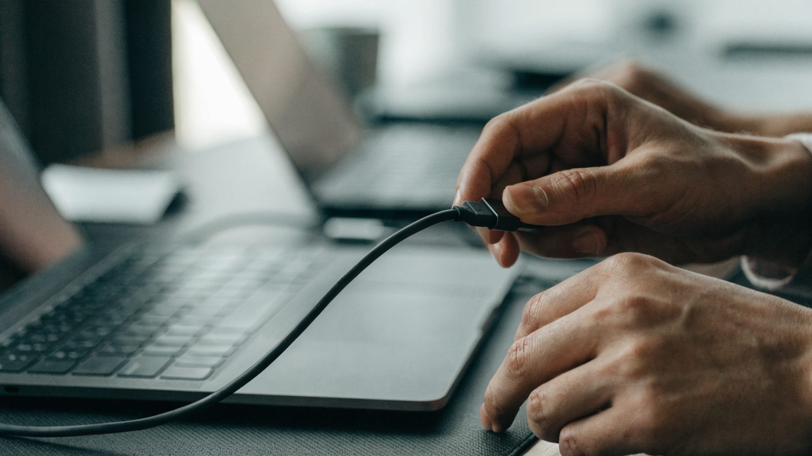 Hands unplugging a charging cable from a slim graphite black laptop on a dark desk mat