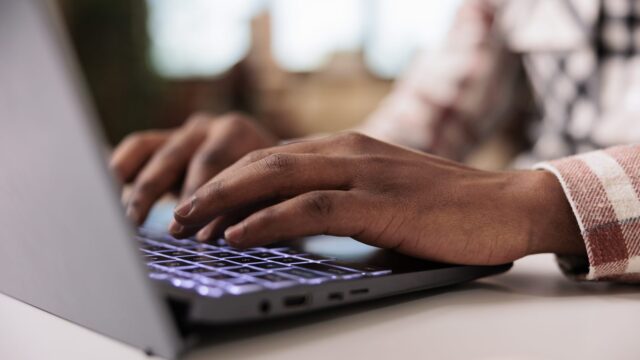 Closeup of male programmer writing code on laptop keyboard while working remote from home living room.…