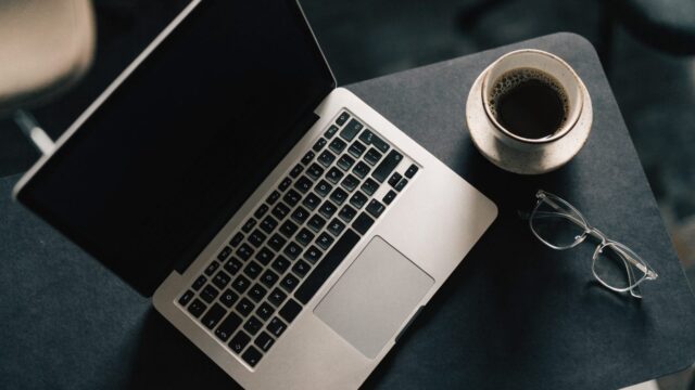 Top-down view of a silver laptop on a dark desk mat with a coffee cup and glasses