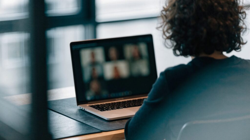A person seen from behind on a video call on a silver laptop on a dark desk mat
