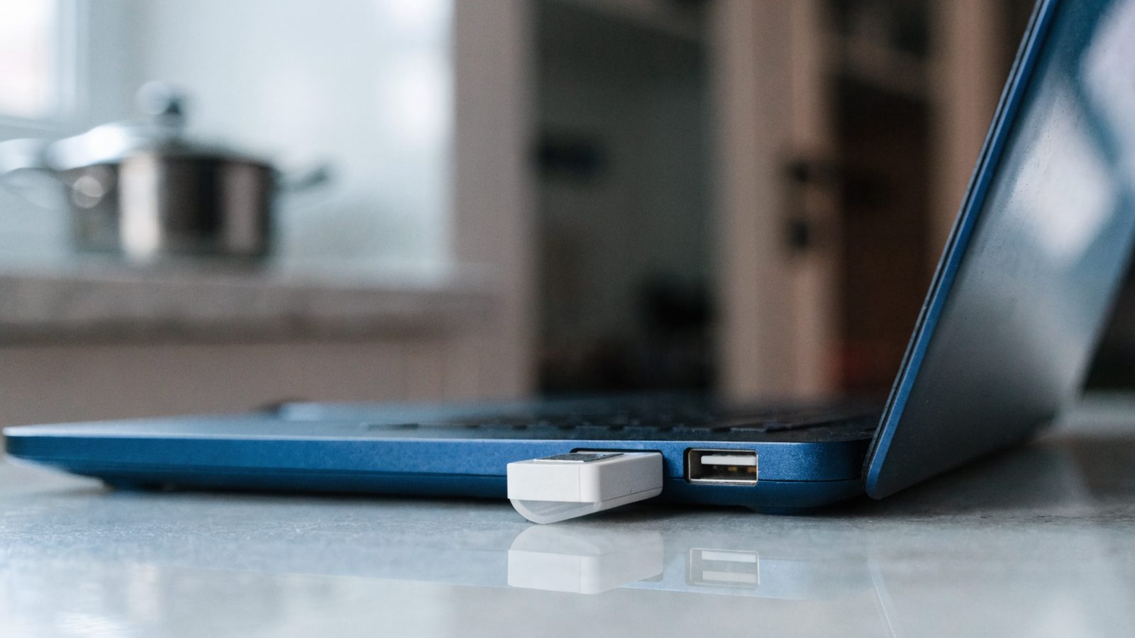 A USB flash drive plugged into the side port of a dark blue laptop on a kitchen table