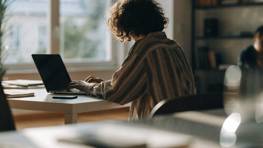 A person seen from behind working on a graphite black laptop in a bright home office