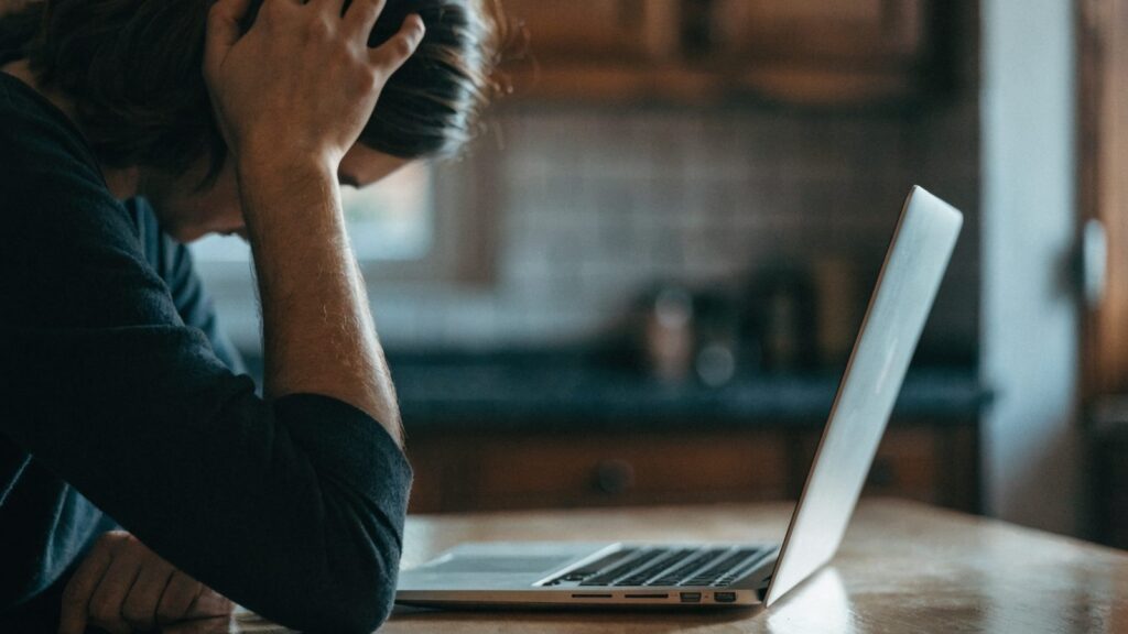 A person resting their forehead on one hand in front of a silver laptop on a kitchen table
