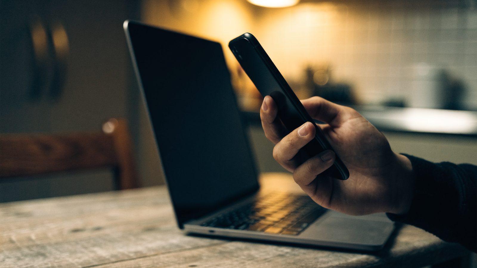A hand holding a smartphone in front of a matte grey laptop on a kitchen table
