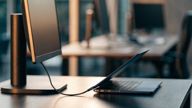 A dark blue laptop connected to a large external monitor with a blank dark screen via a cable on an office des