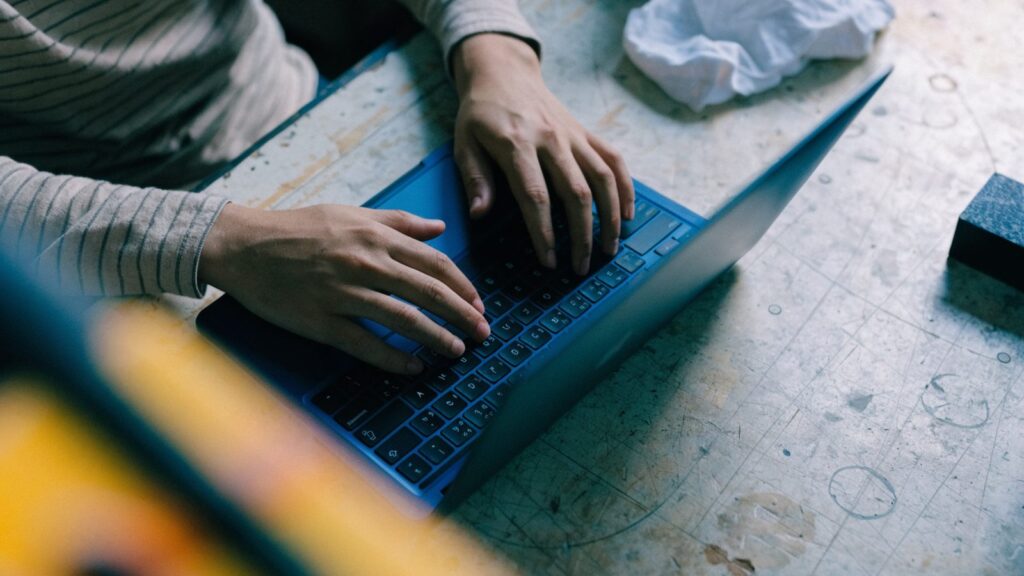 Hands typing on the keyboard of a dark blue laptop on a workbench