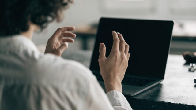 A person seen from behind with hands raised at a matte grey laptop with a dark blank screen on a workbench