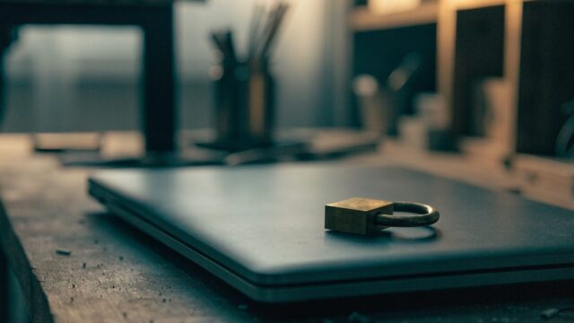 A small brass padlock resting on the closed lid of a graphite black laptop on a workbench