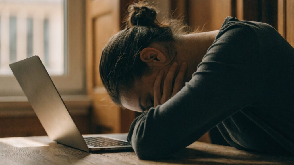 A person resting their forehead on one hand in front of a matte grey laptop on a wooden desk