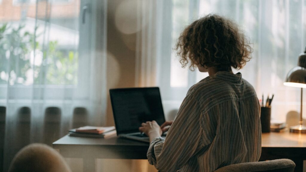 A person seen from behind working on a matte grey laptop in a bright home office