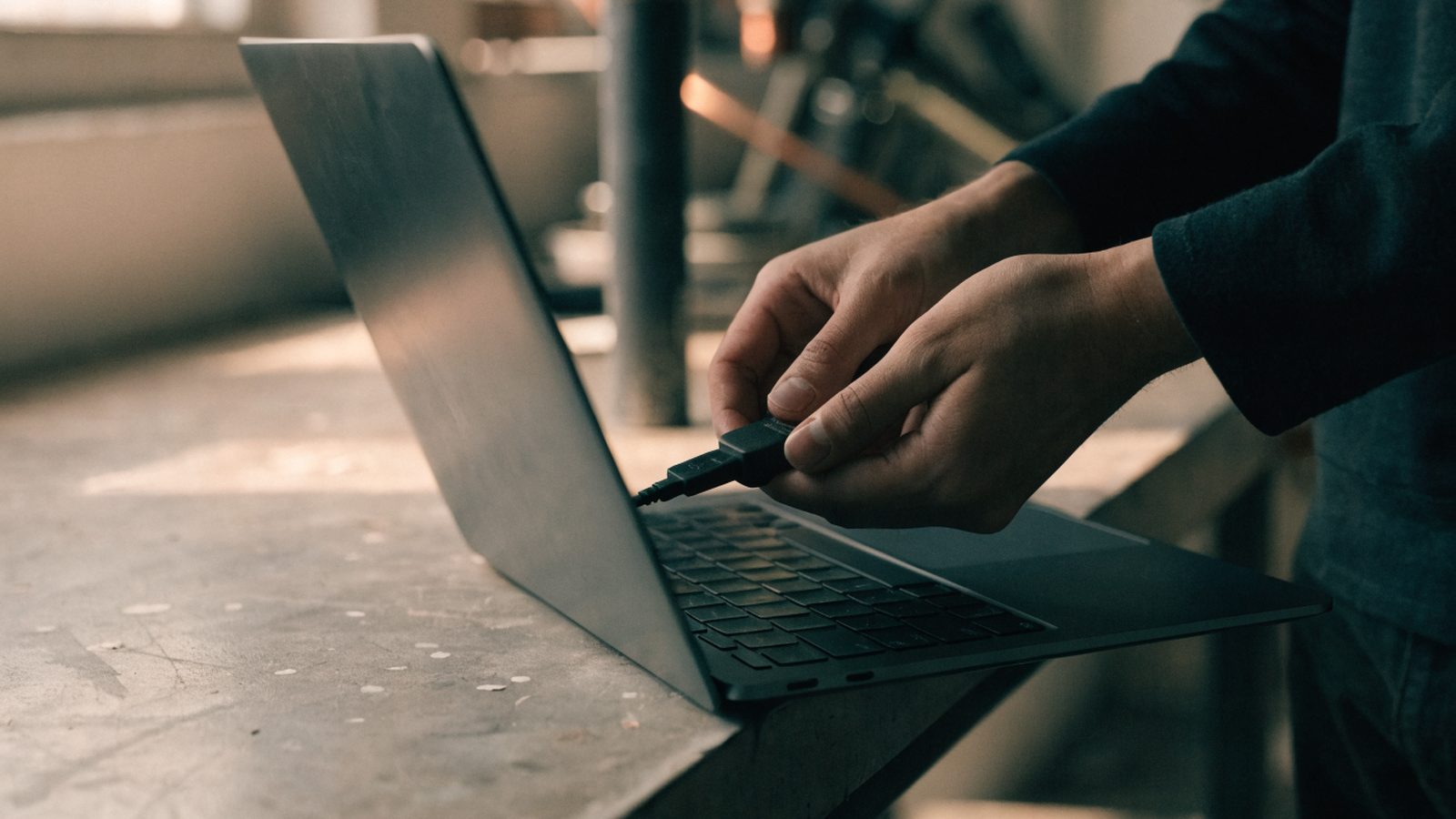 Hands unplugging a charging cable from a slim graphite black laptop on a workbench