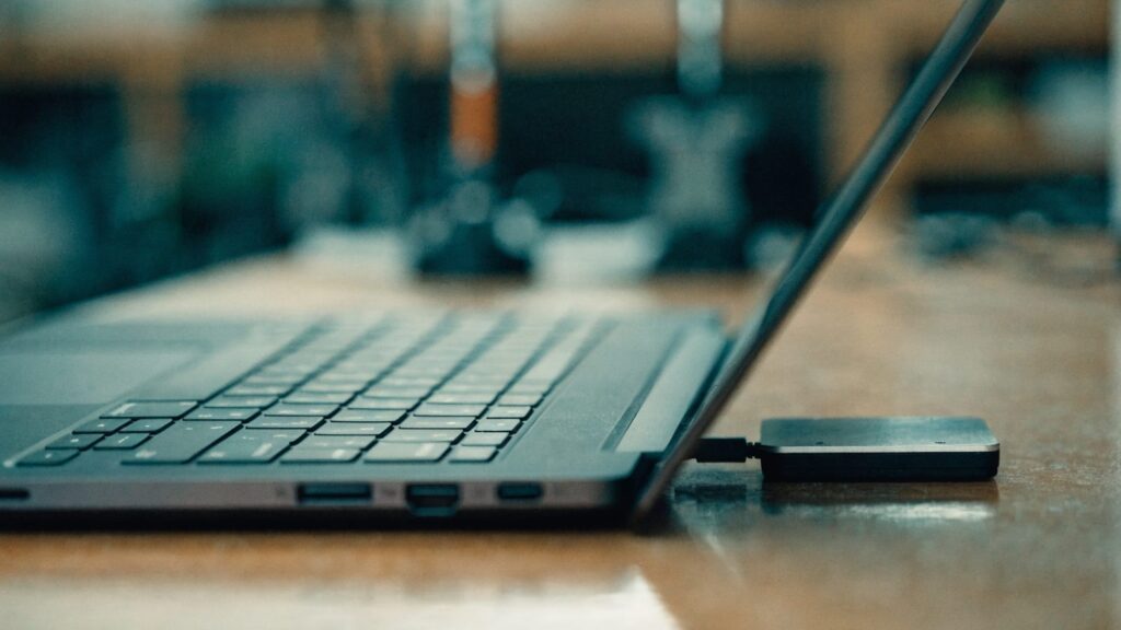 A small external hard drive connected by cable to a graphite black laptop on a workbench