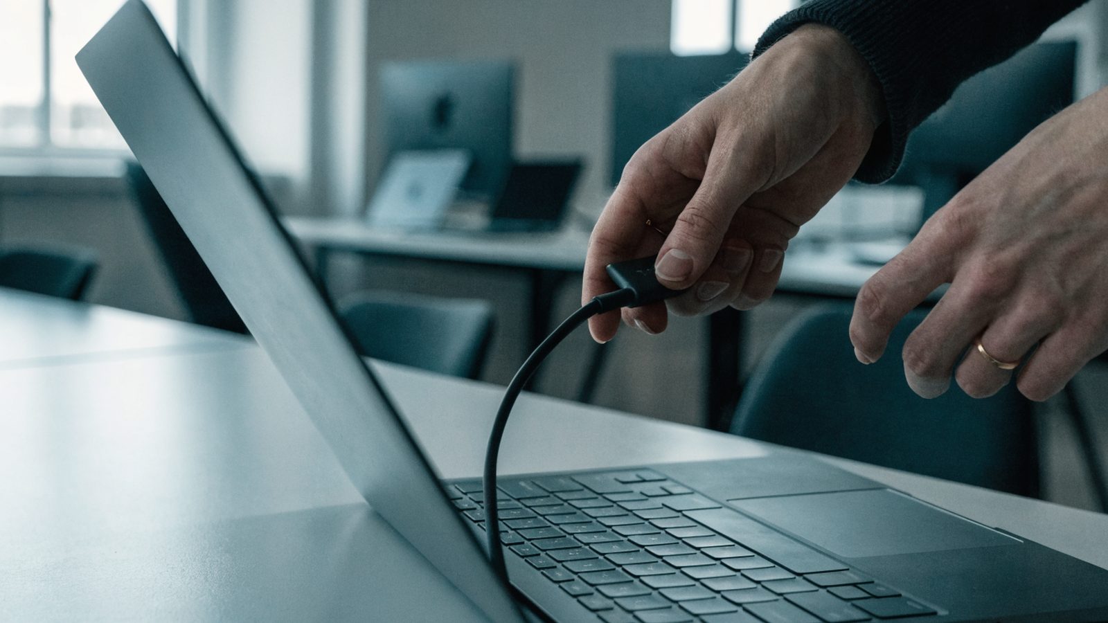 Hands unplugging a charging cable from a slim graphite black laptop on an office desk