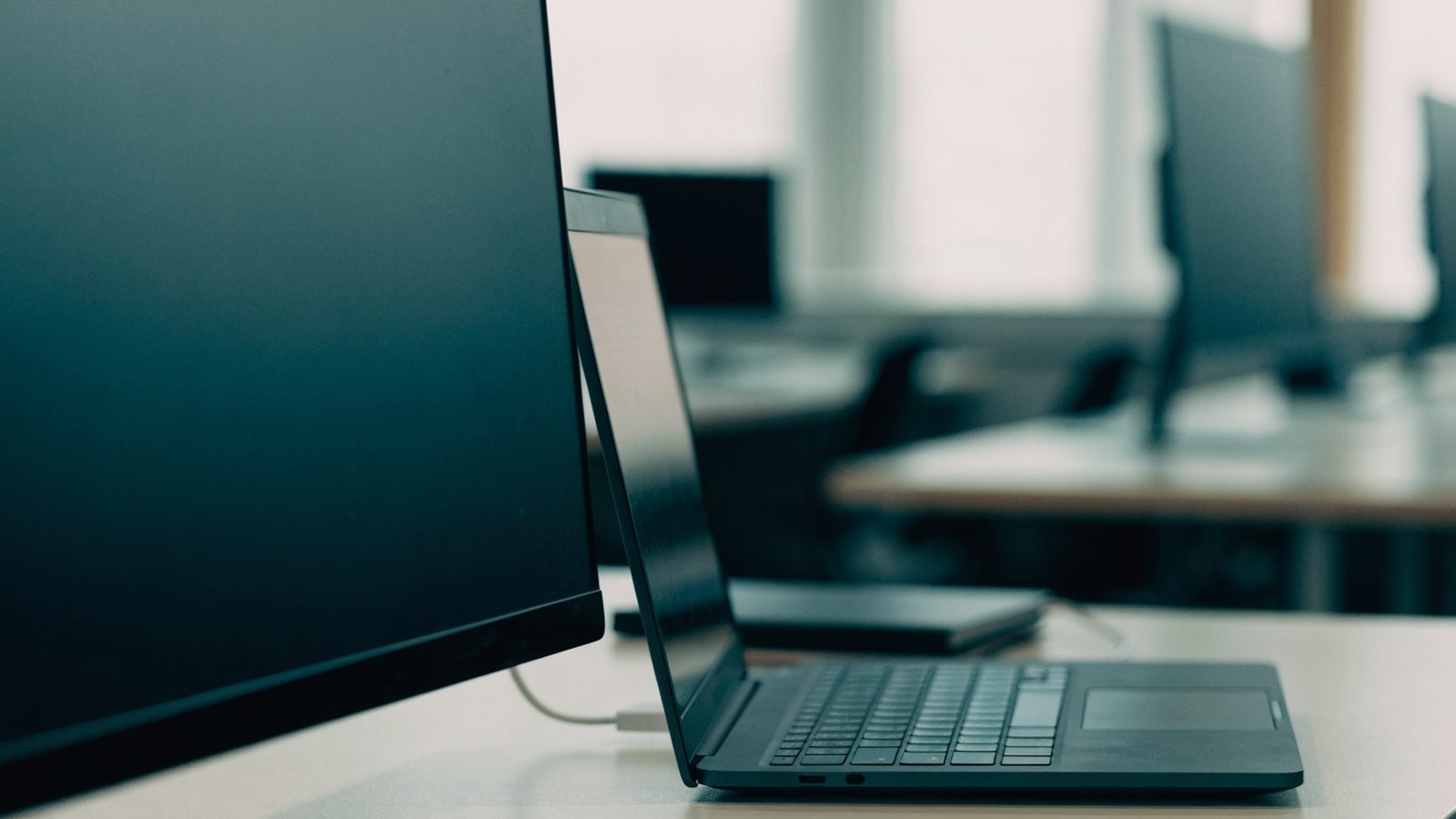 A graphite black laptop connected to a large external monitor with a blank dark screen via a cable on an offic