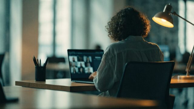 A person seen from behind on a video call on a dark blue laptop on an office desk