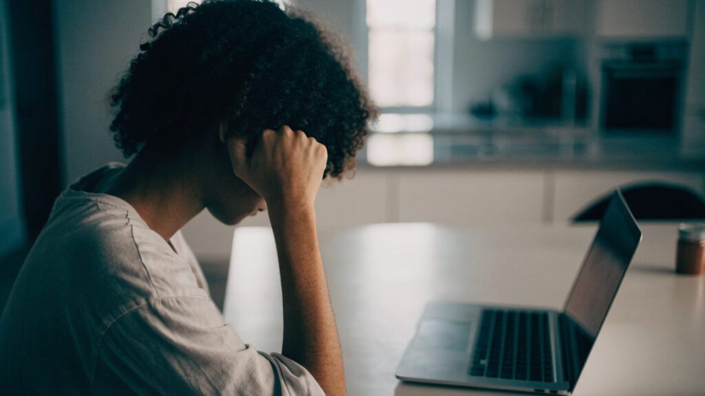 A person resting their forehead on one hand in front of a silver laptop on a kitchen table