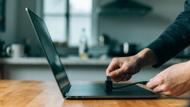 Hands unplugging a charging cable from a slim graphite black laptop on a kitchen table
