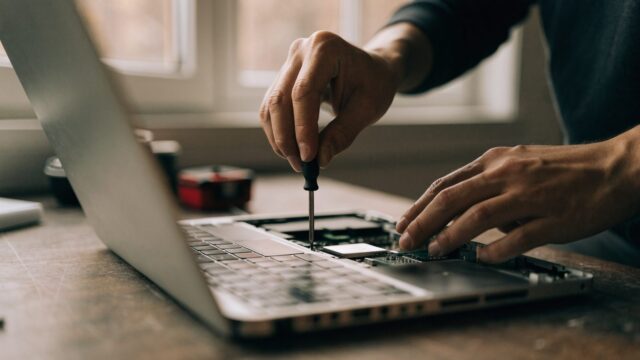 A technician's hands holding a small screwdriver over an opened silver laptop with internal components visible