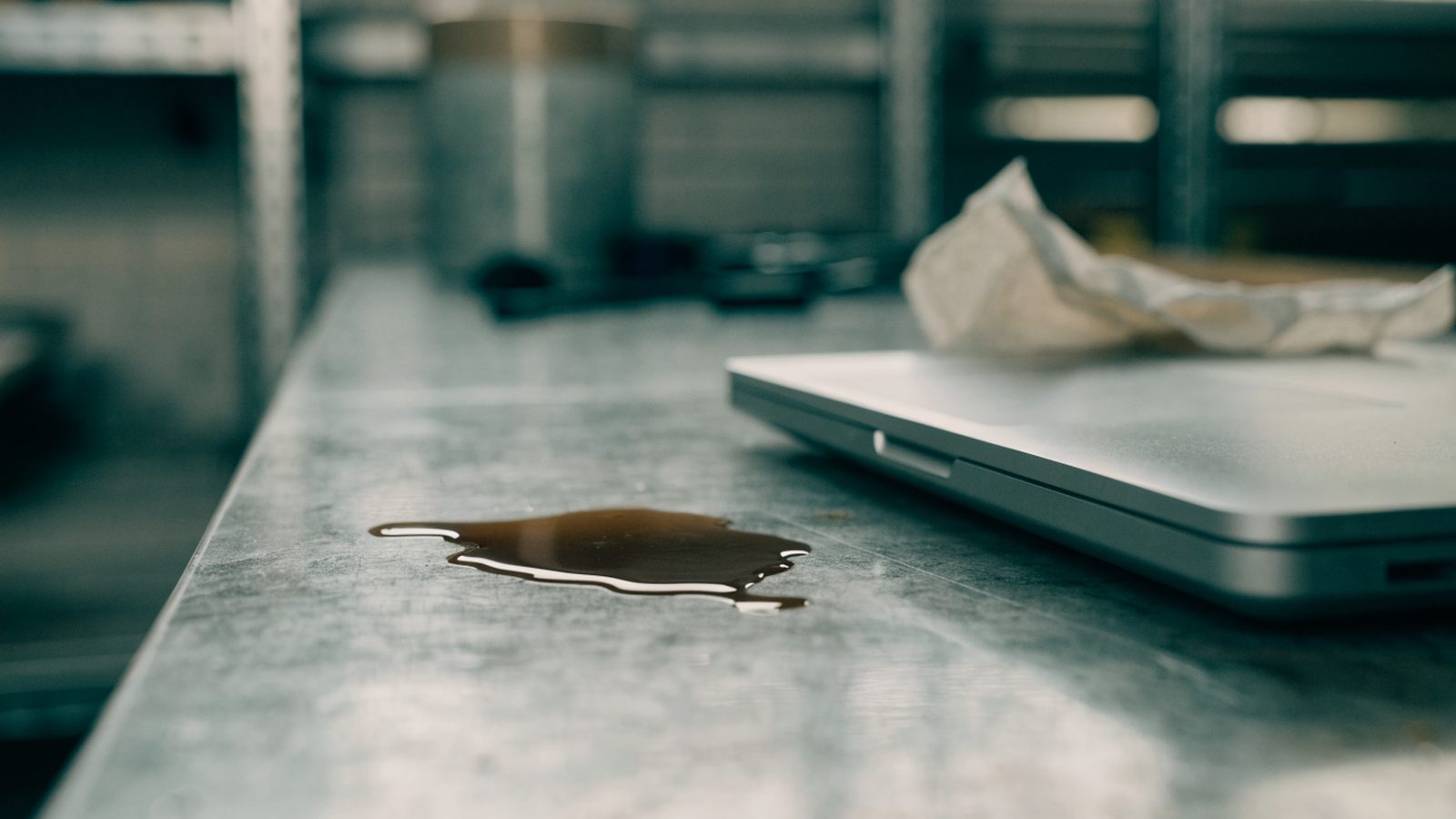A small puddle of spilled coffee next to a matte grey laptop on a workbench