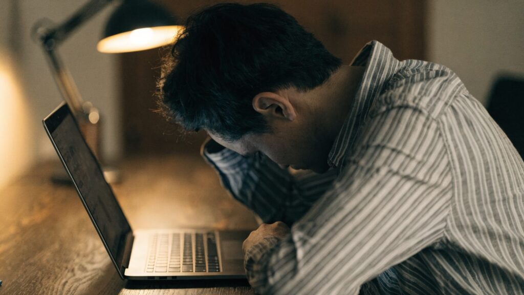 A person resting their forehead on one hand in front of a graphite black laptop on a wooden desk