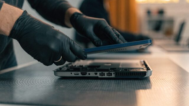 A technician lifting the bottom cover off a dark blue laptop on an anti-static mat