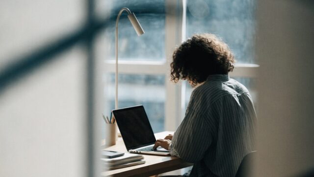 A person seen from behind working on a silver laptop in a bright home office
