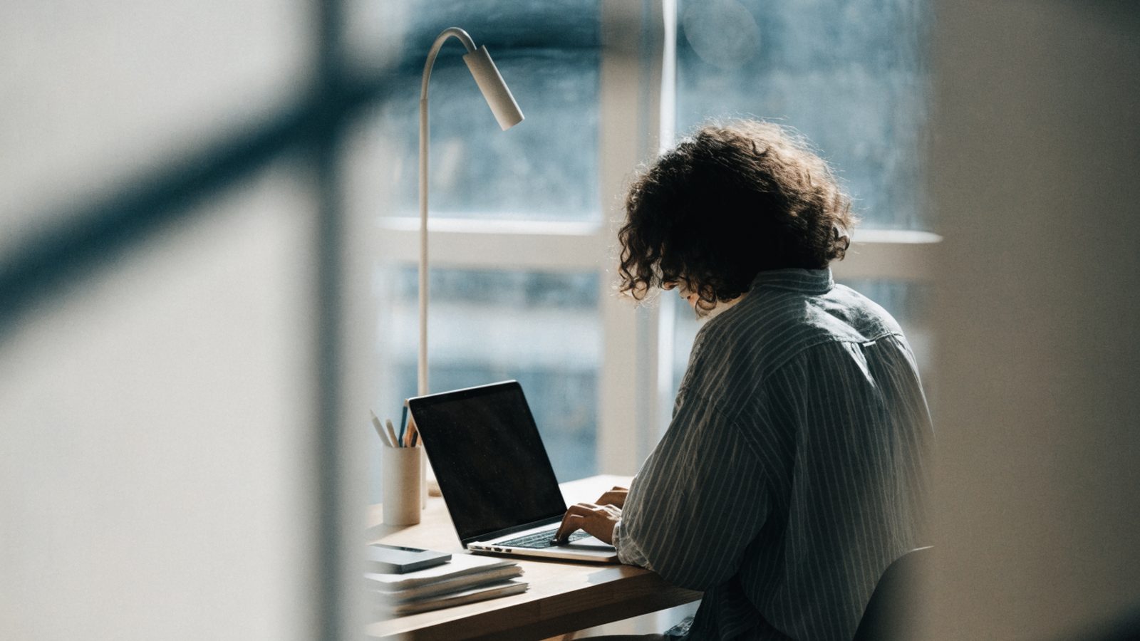 A person seen from behind working on a silver laptop in a bright home office
