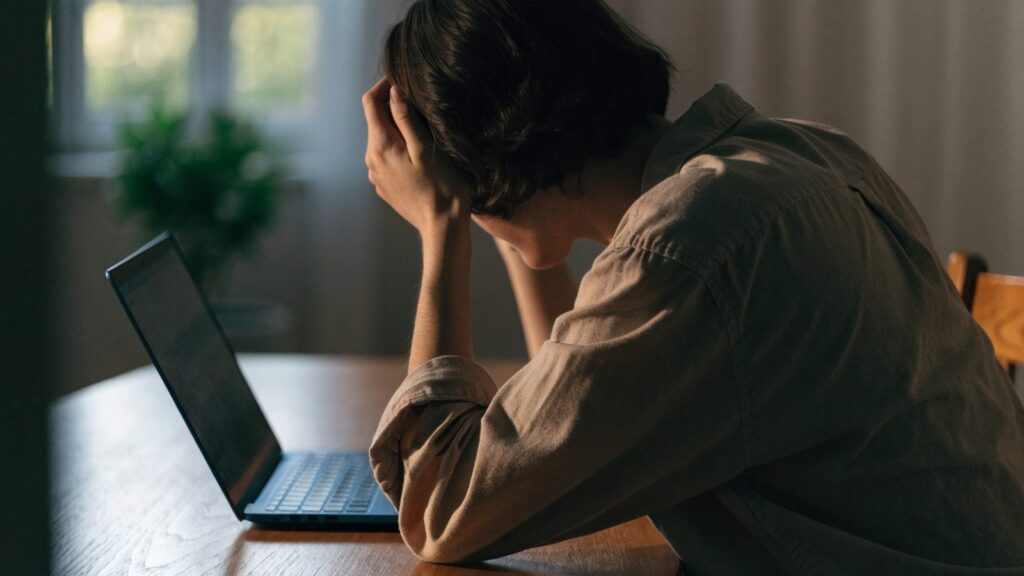 A person resting their forehead on one hand in front of a dark blue laptop on a wooden desk
