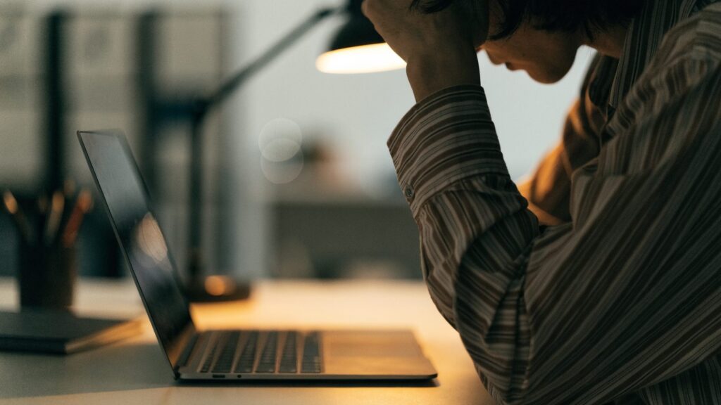 A person resting their forehead on one hand in front of a graphite black laptop on an office desk