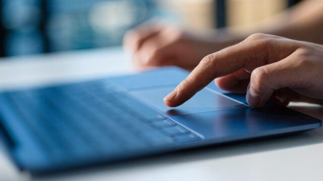 A finger gliding on the touchpad of a dark blue laptop on a white desk