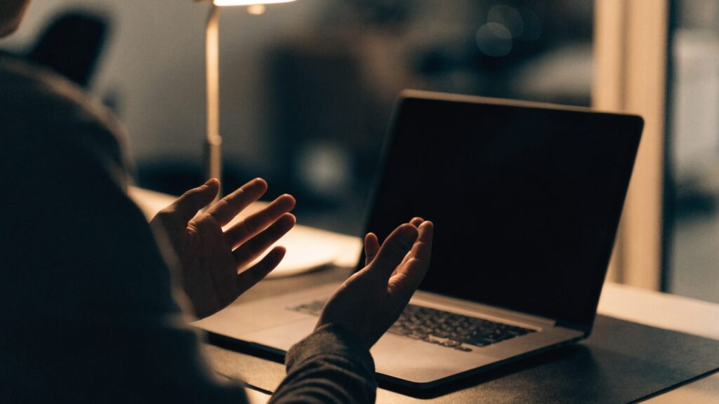 A person seen from behind with hands raised at a matte grey laptop with a dark blank screen on a dark desk mat