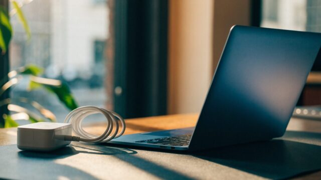 A dark blue laptop on a dark desk mat next to a compact white power adapter with its cable neatly coiled