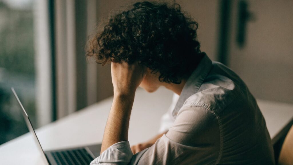 A person resting their forehead on one hand in front of a matte grey laptop on a white desk