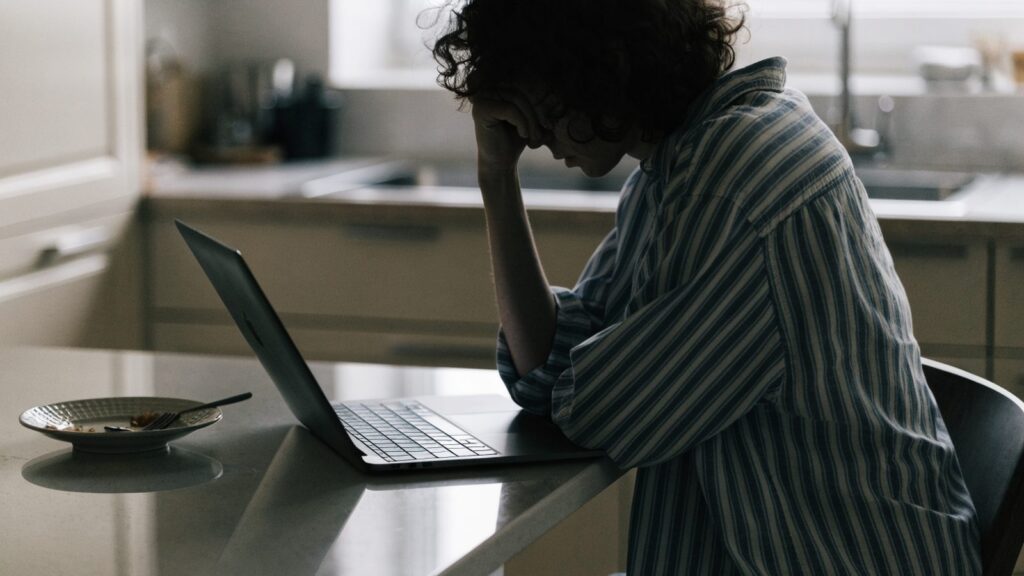 A person resting their forehead on one hand in front of a graphite black laptop on a kitchen table
