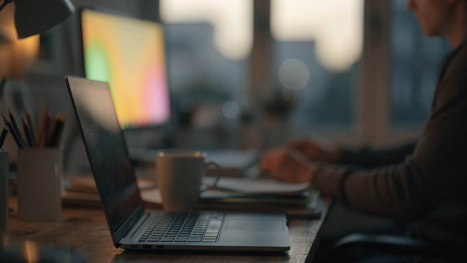 A developer's desk at night with a dark blue laptop and a second monitor showing soft blurred colorful glow