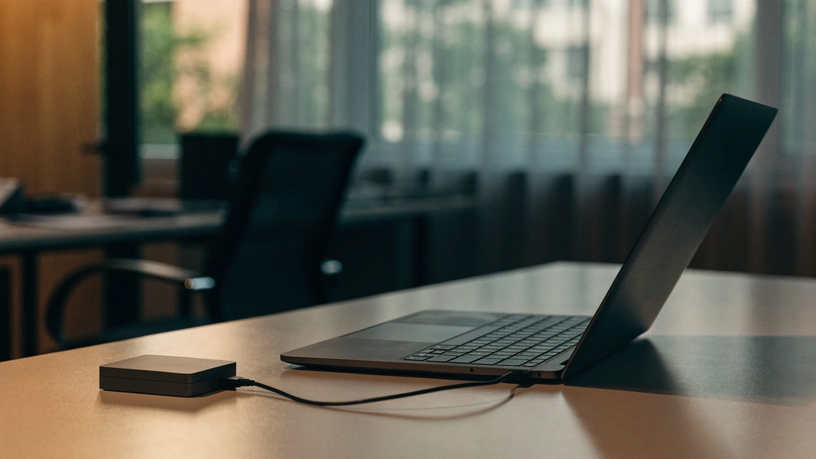 A small external hard drive connected by cable to a graphite black laptop on an office desk