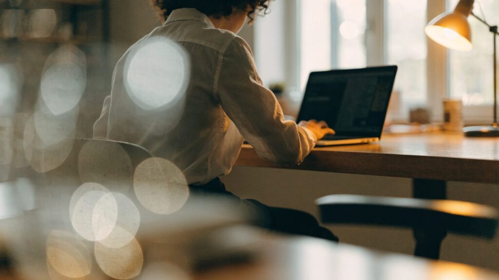 A person seen from behind working on a silver laptop in a bright home office