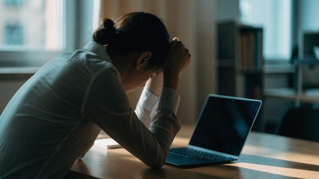 A person resting their forehead on one hand in front of a dark blue laptop on an office desk